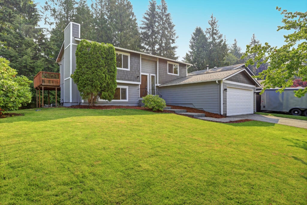 A two-story gray house with white trim, a double garage, and a well-maintained lawn is surrounded by trees and shrubs on a sunny day. Featuring quality roofing, this Lorton home also boasts a wooden deck on the left side.
