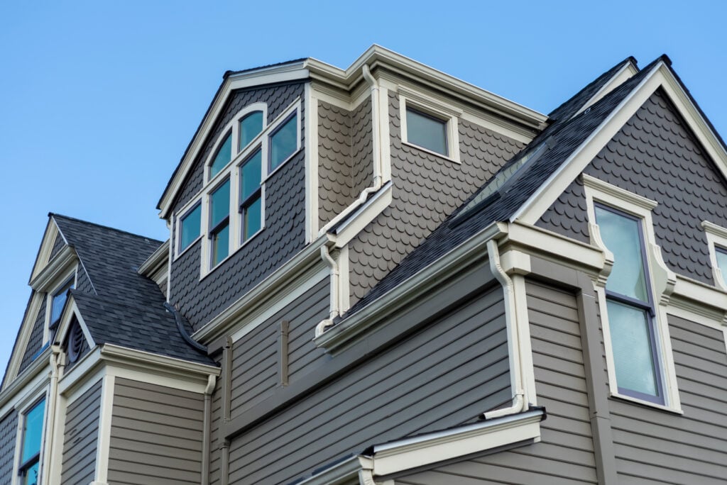 Gray two-story house with white trim, multiple large windows, and gabled roofs—a perfect example of stylish roofing Falls Church homeowners admire. The exterior features vertical and fish-scale siding designs, all set against a clear blue sky.