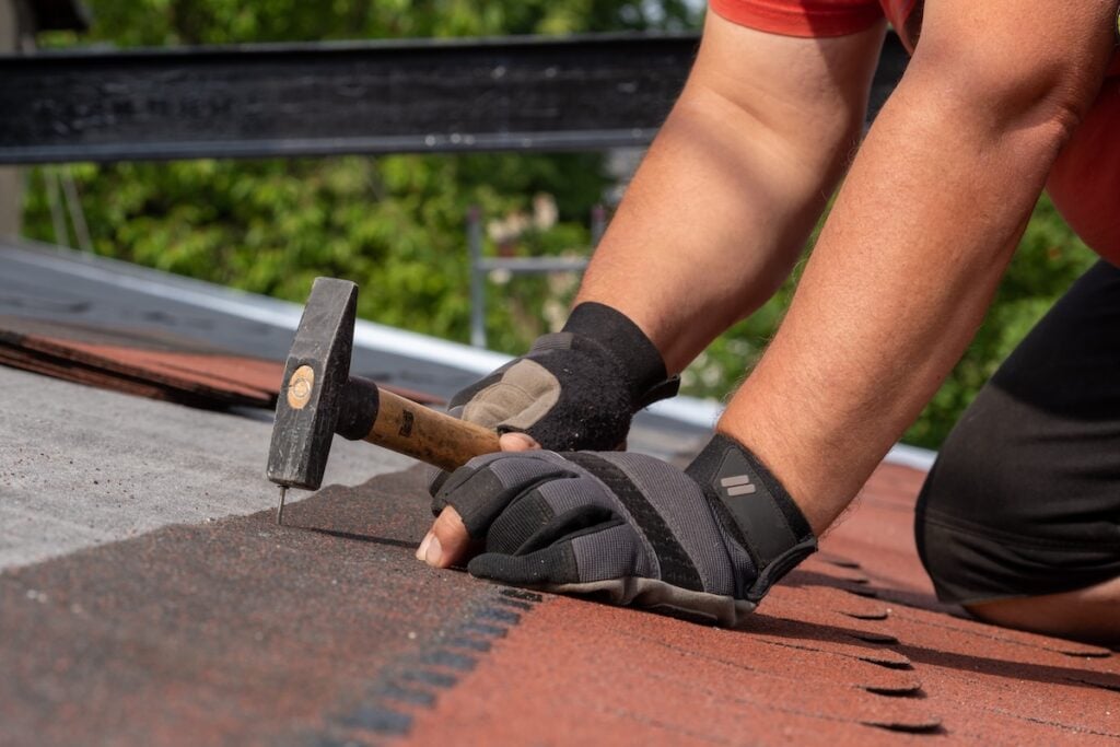Person wearing gloves uses a hammer to nail down asphalt shingles on a roof, working with both hands close to the surface. Trees and metal scaffolding are visible in the blurred background.