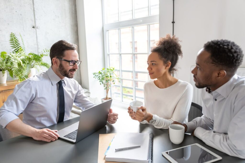Three people sit at a table in a bright office, discussing a roofing Vienna VA project. One man uses a laptop, while the others hold coffee mugs. A notepad and pen are on the table, with large windows and plants in the background.