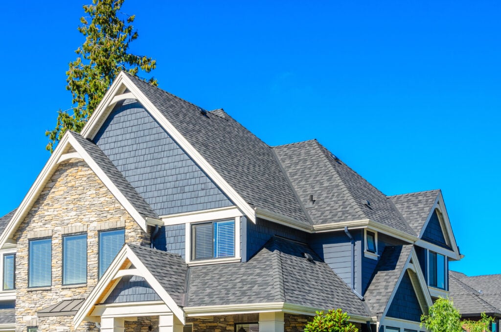 A two-story house with gray shingles, blue siding, stone details, and a steeply pitched roof—an ideal example of expert roofing Falls Church residents admire—set against a clear blue sky and framed by green trees.