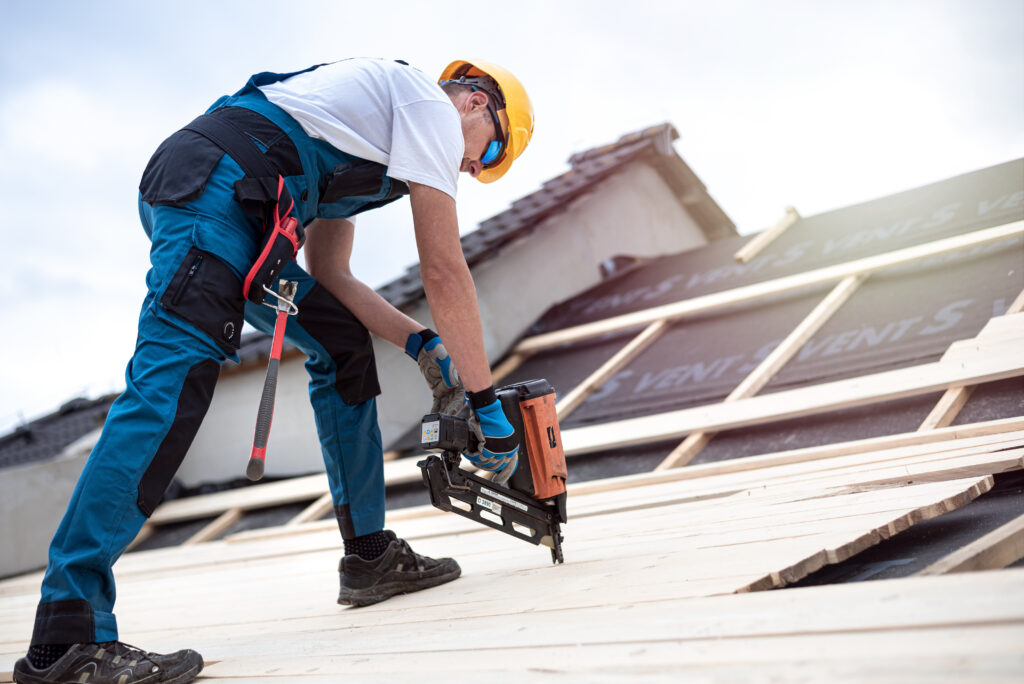 A construction worker in safety gear uses a nail gun to secure wooden boards on a sloped roofing Falls Church project, with a partly cloudy sky in the background.