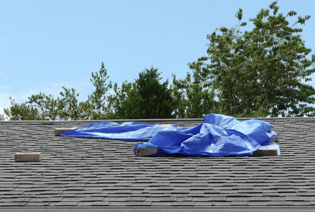 A blue tarp held down by bricks covers part of a shingled roof, possibly awaiting roofing Fairfax repairs. Green trees are visible in the background under a clear sky.