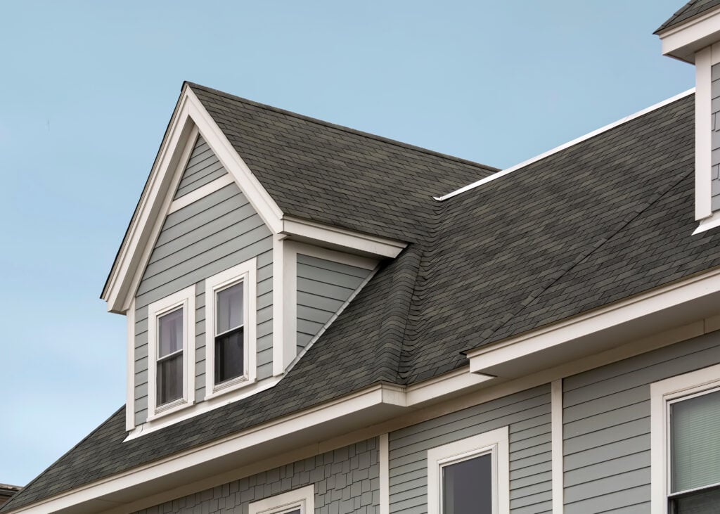 A close-up view of the upper exterior of a house with gray siding, white trim, and dark gray roof shingles, showcasing expert roofing Falls Church craftsmanship against a clear blue sky. Two windows are visible on the gable end.