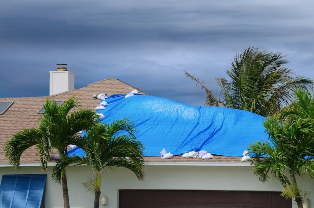 A blue tarp secured with sandbags covers part of a house roof, likely damaged by recent storms—roofing Falls Church services may be needed. Palm trees stand nearby under a cloudy, stormy sky.