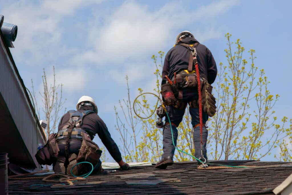 Two workers wearing safety harnesses and helmets are repairing or installing shingles on a sloped roof in Vienna, VA, with safety ropes attached; trees and blue sky are visible in the background. Perfect example of skilled roofing Vienna VA services.