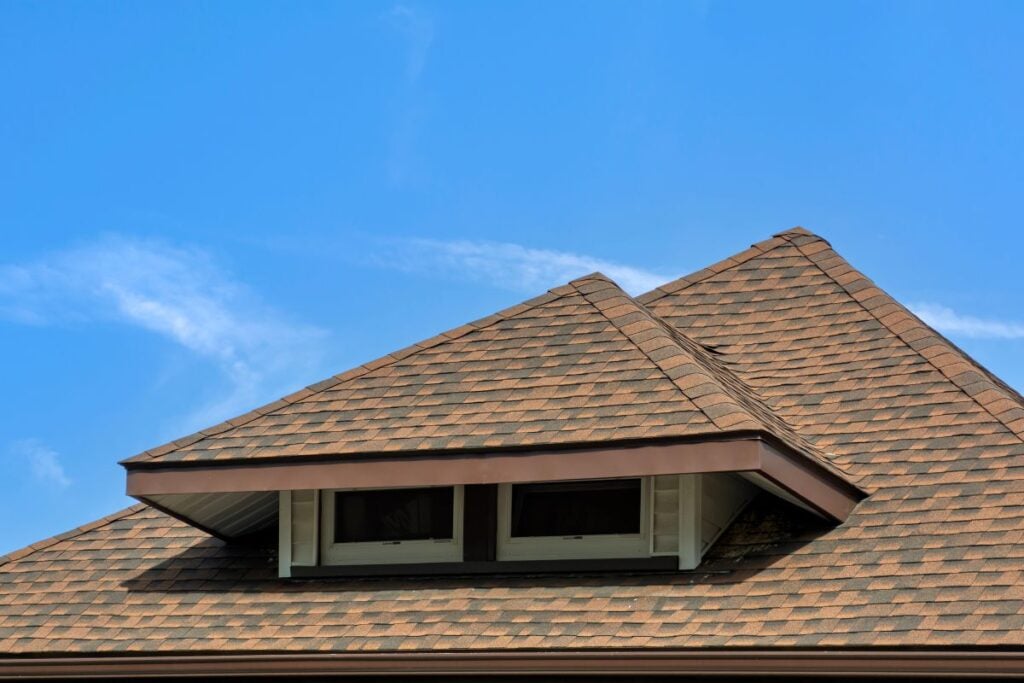 Roof Replacement Cost Falls Church Close-up of a house roof with brown asphalt shingles, featuring two small windows under a dormer, set against a clear blue sky with a few wispy clouds.