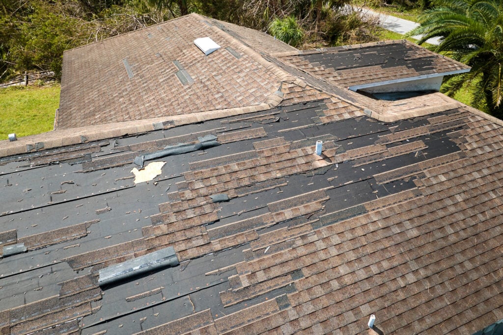 A house roof in disrepair, likely needing roofing Lorton services, with many missing and damaged shingles exposing the black underlayment. Scattered shingles suggest storm damage. Trees and a driveway can be seen in the background.