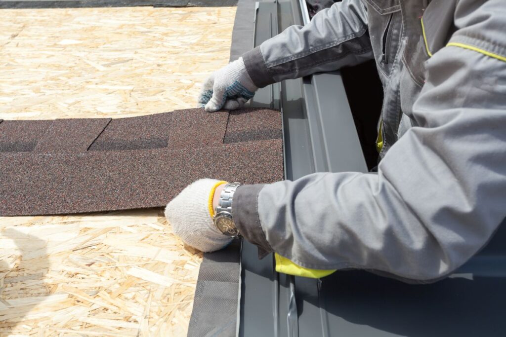 A person wearing work gloves and a gray jacket is installing an asphalt shingle onto a wooden roof structure, preparing for roofing work in Vienna, VA.