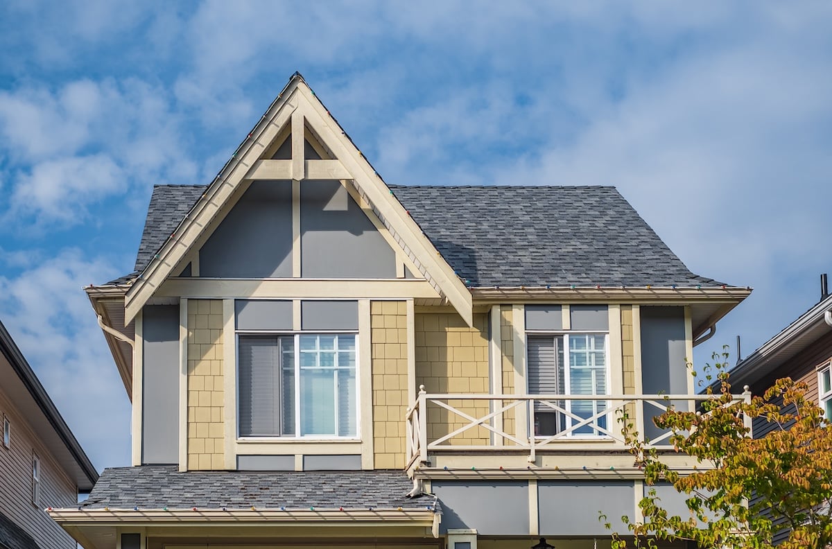 A two-story house with a steep gray roof, beige and gray exterior, white-framed windows, and a small balcony, set against a partly cloudy blue sky.