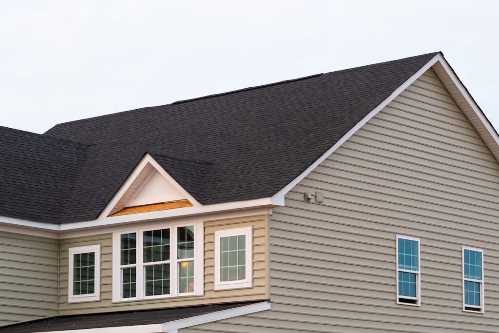 The image shows the upper portion of a beige house with horizontal siding, several white-trimmed windows, and a dark shingled roof against a light sky background.