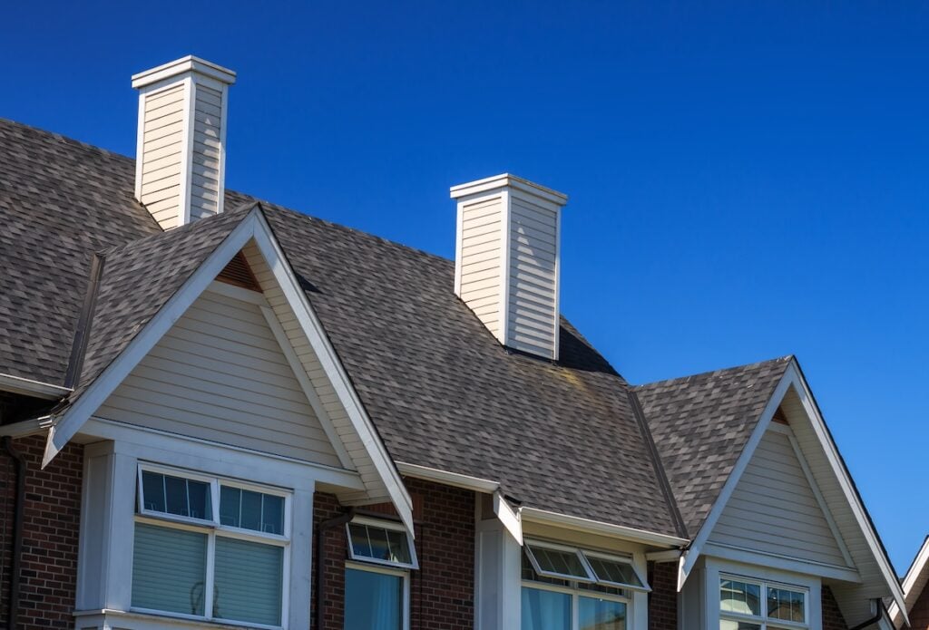 Three house rooftops with gray shingles, white chimneys, and triangular gables under a clear blue sky. The homes have brick siding on the lower level and white trim around the windows and roof.