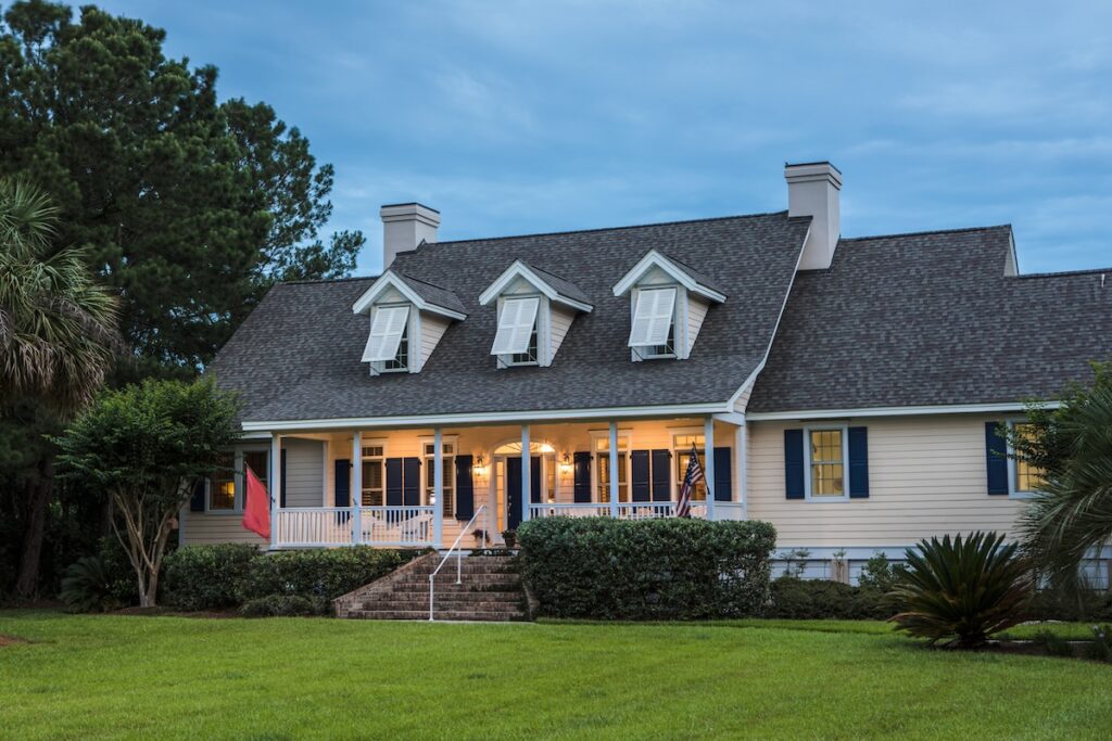 A two-story suburban house with a large front porch, dormer windows, and a well-kept lawn. The porch is lit, featuring steps, railings, and American and red flags. Trees and shrubs surround the home.