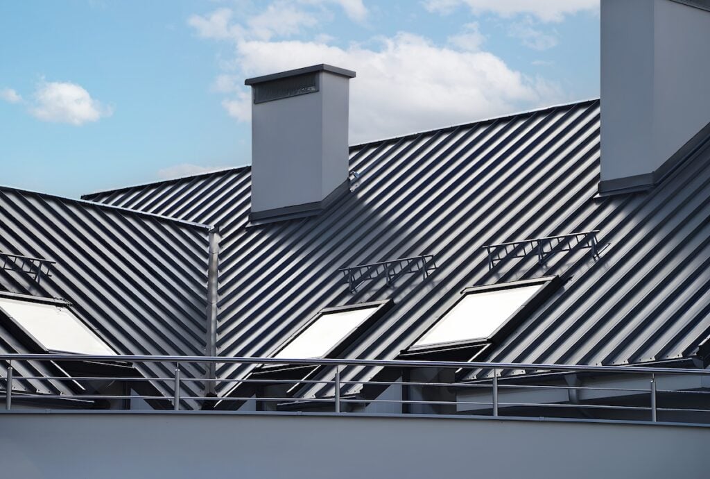 A modern building with a dark metal sloped roof, three large skylights, and two white chimneys, set against a blue sky with clouds. The foreground shows a metal railing.