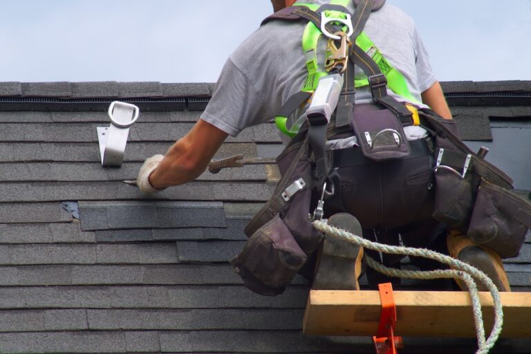 A worker wearing a safety harness and tool belt repairs gray roof shingles while kneeling on a wooden platform. Nearby, a vent pipe and various tools are visible on the roof.