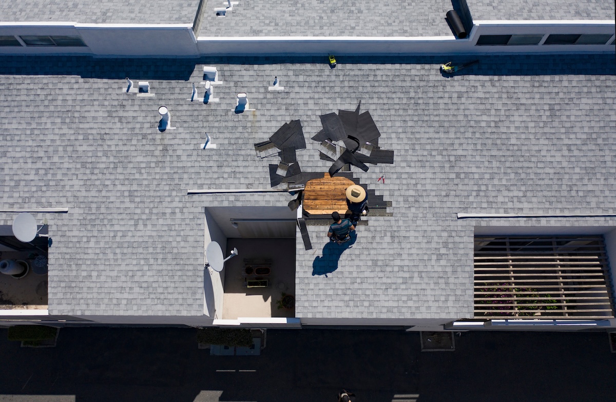 Aerial view of a person repairing a gray shingled roof. Roofing materials are spread nearby, and the person is standing on a wooden platform wearing a hat. The sunlight casts shadows on the roof.