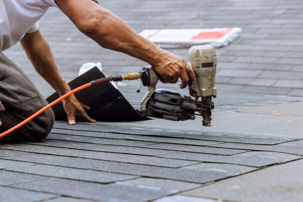 roof replacement cost arlington A person uses a pneumatic nail gun to secure asphalt shingles to a roof during a roofing installation or repair. The individual is kneeling and positioning the shingles carefully.