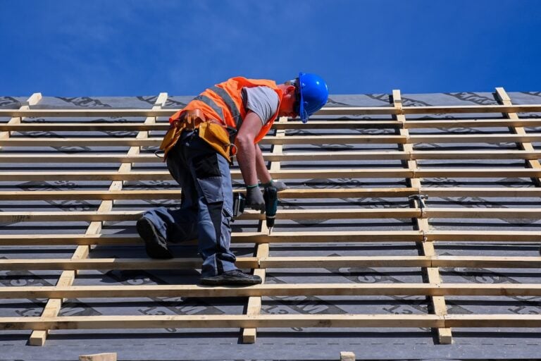 A construction worker in a blue hard hat and orange safety vest is installing wooden battens on a sloped roof under a clear blue sky. The worker is using a power tool while balancing on the roof structure.