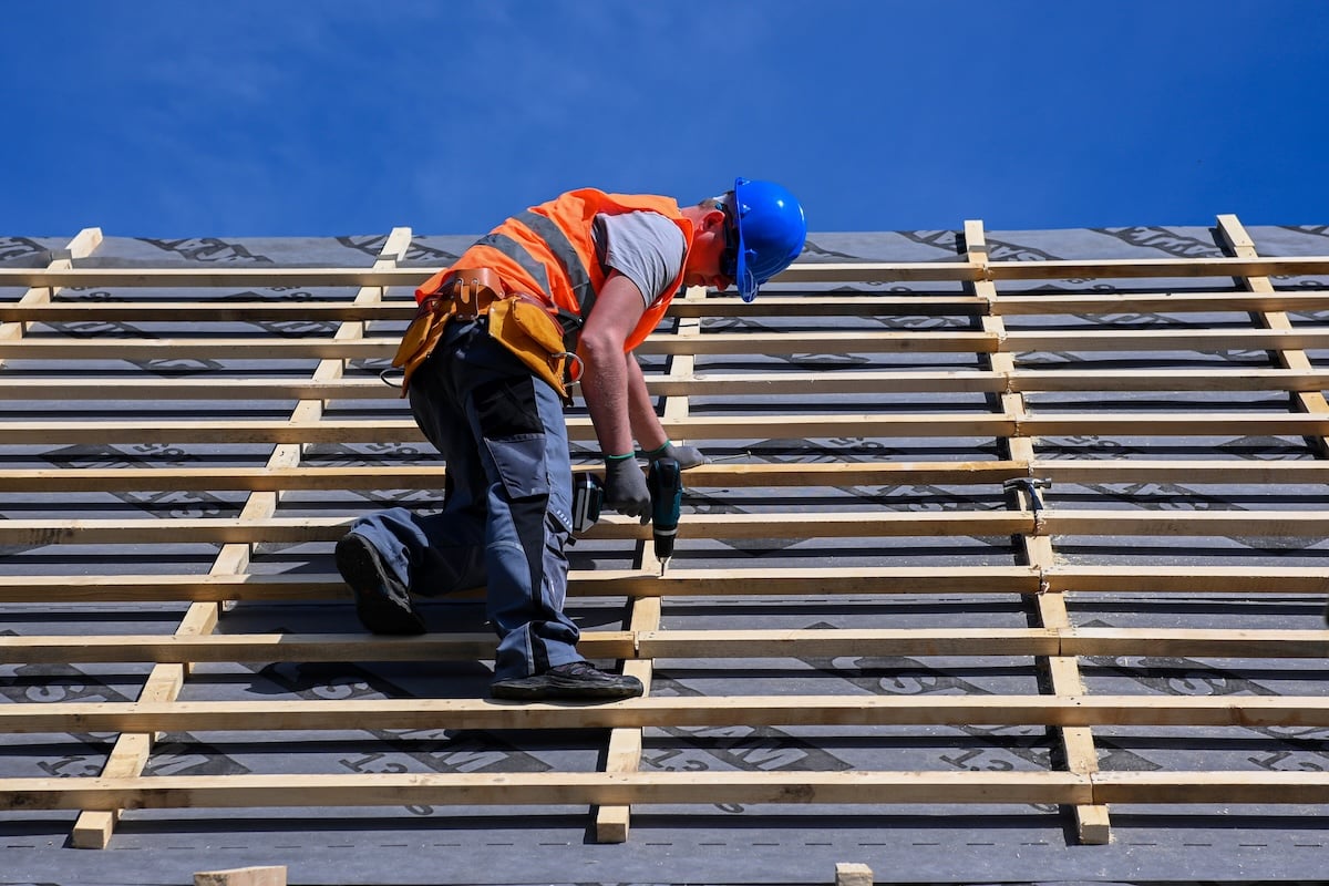 A construction worker in a blue hard hat and orange safety vest is installing wooden battens on a sloped roof under a clear blue sky. The worker is using a power tool while balancing on the roof structure.