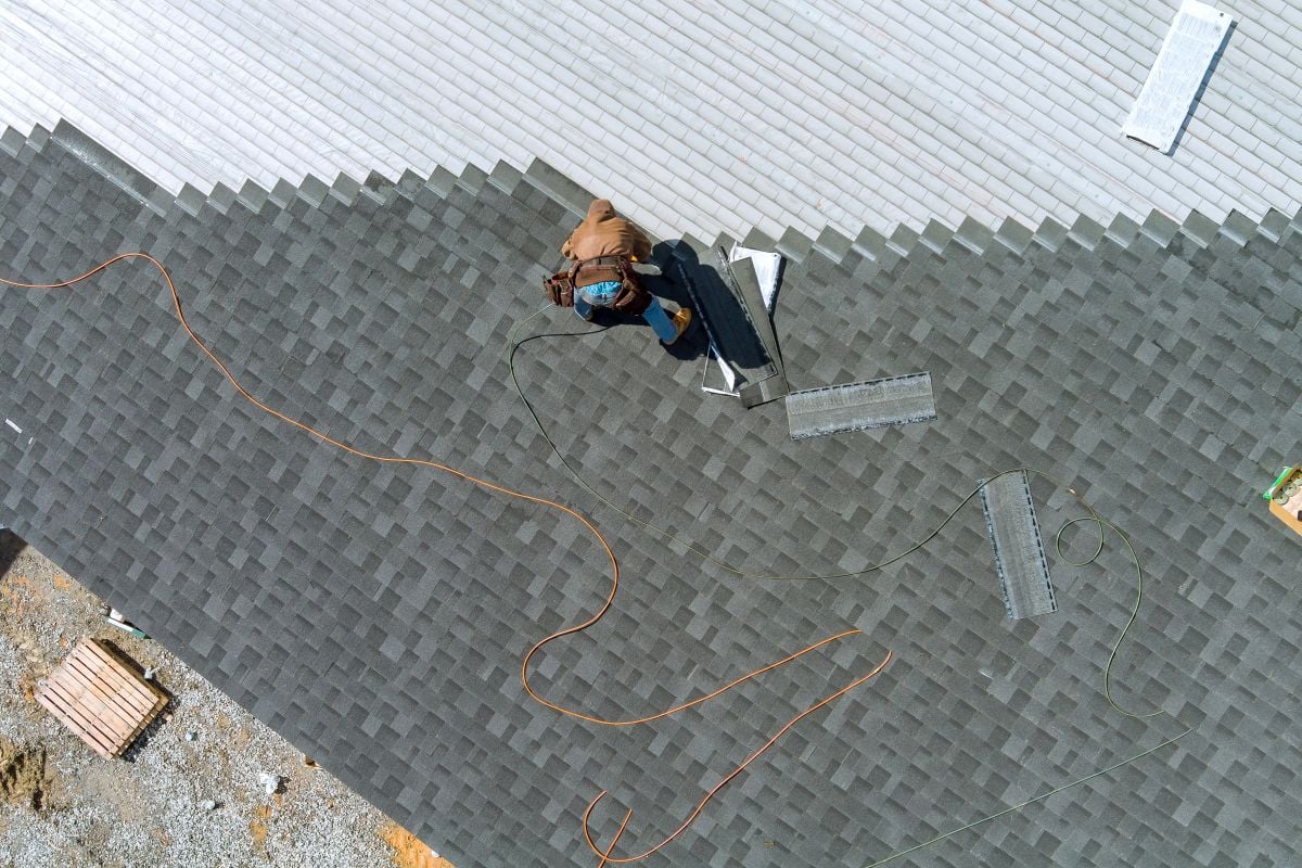 A worker installs asphalt shingles on a roof, with tools and materials nearby. The roof is partly finished, showing a clear line between completed and unfinished sections. An orange extension cord is visible.