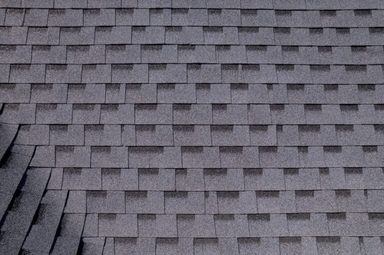 Close-up view of overlapping gray asphalt roof shingles arranged in a neat, repetitive pattern, with a small section on the lower left appearing slightly uneven.