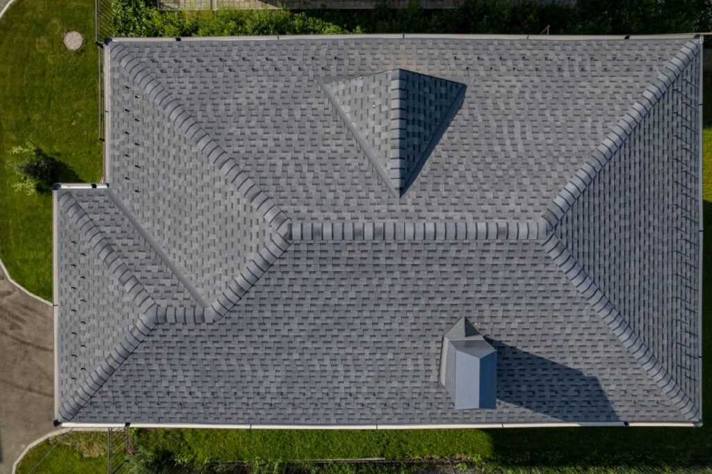 Aerial view of a house roof with gray shingles, featuring multiple ridges and a small vent structure, surrounded by green grass and some bushes along the edge.