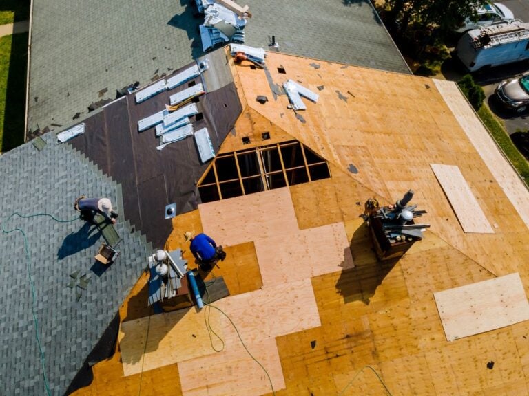 Aerial view of roofers installing new plywood sheathing and shingles on a house roof, with roofing materials and tools scattered around. Some areas are covered with new shingles, others with exposed wood.