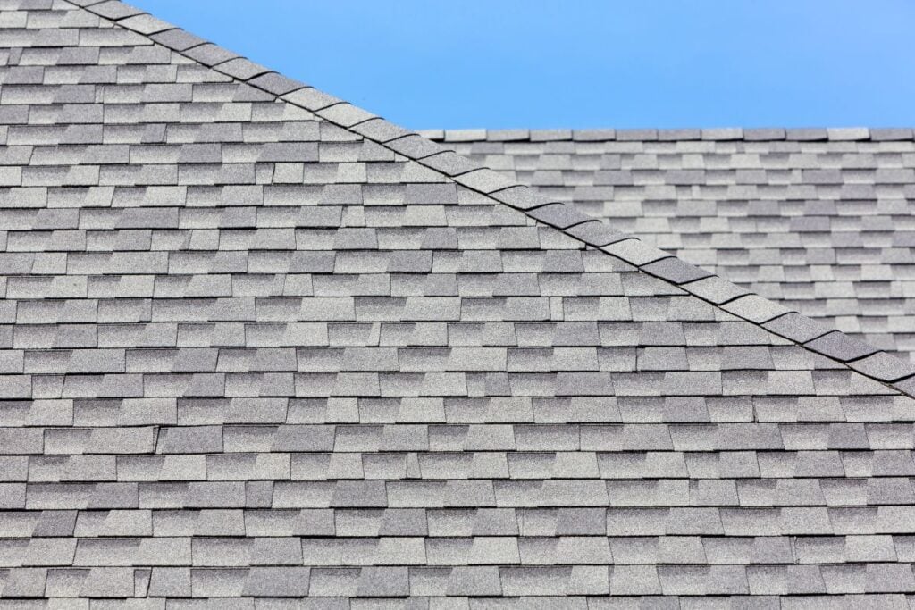 Close-up view of two intersecting gray asphalt shingle rooftops against a clear blue sky. The rooftops form a diagonal line where they meet, showing the texture and pattern of the shingles.