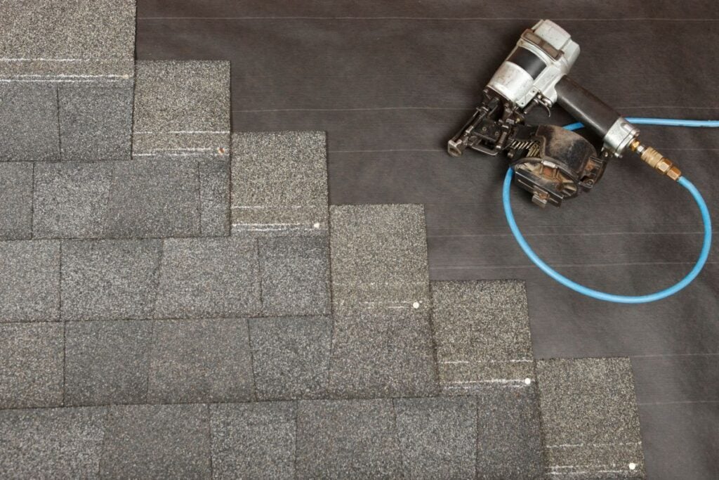Gray asphalt shingles being installed on a roof with a nail gun and blue air hose resting nearby on black underlayment. The shingles are arranged in a stepped pattern.