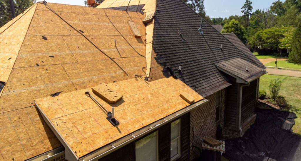 A house undergoing roof replacement; one section has new plywood sheathing while another section still has old asphalt shingles. Roofing tools and materials are visible, and tarps cover parts of the ground below.