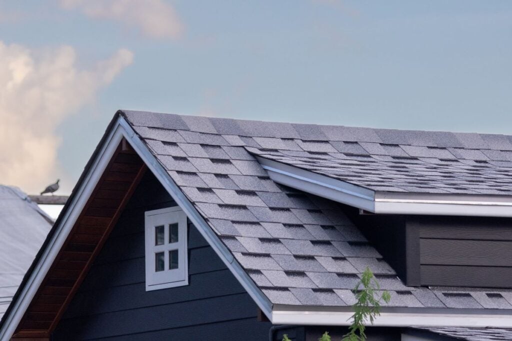 Close-up of a modern house roof with gray asphalt shingles, a small window near the peak, clear blue sky, and a bird perched on a distant rooftop in the background.