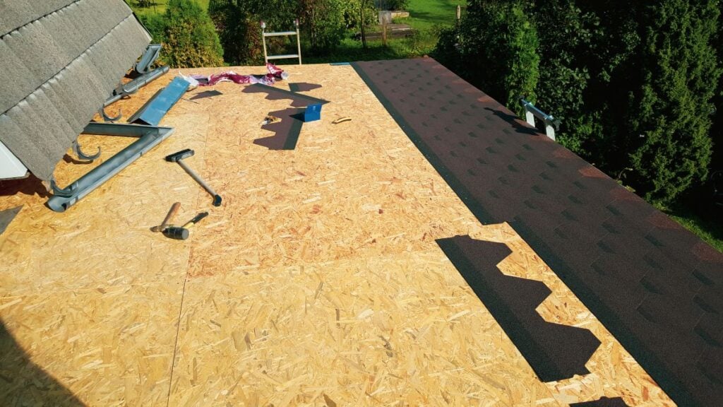 Partially completed roof with wooden sheathing; brown asphalt shingles are being installed, with tools like a hammer and utility knife scattered on the surface. Trees and a backyard are visible in the background.