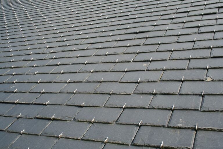 Close-up view of a rooftop covered with overlapping rectangular slate tiles, each secured with visible metal hooks or fasteners, creating a uniform, textured pattern across the surface.
