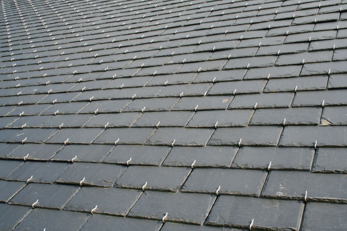 Close-up view of a rooftop covered with overlapping rectangular slate tiles, each secured with visible metal hooks or fasteners, creating a uniform, textured pattern across the surface.