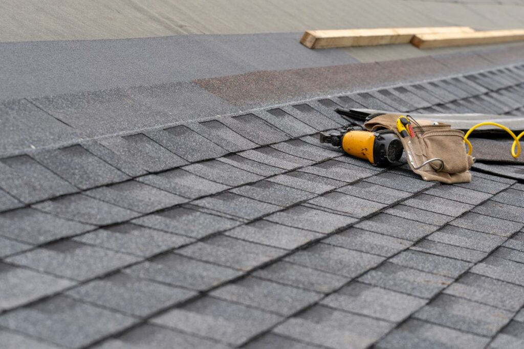 Close-up of gray asphalt roof shingles being installed, with a pneumatic nail gun, tool belt, and wooden boards resting on the roof.