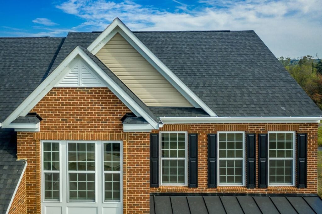 Close-up of a brick house exterior with black shingle roofing, white-framed windows, black shutters, and beige siding under the gable. The sky is partly cloudy in the background.