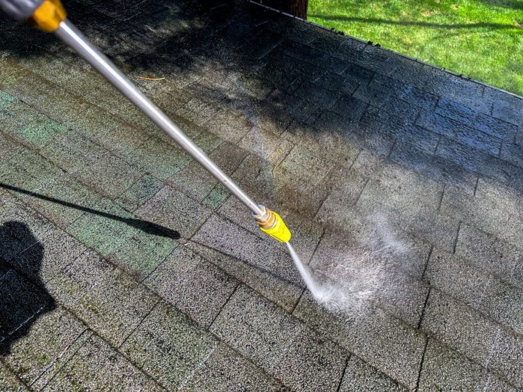 A close-up of a pressure washer cleaning a shingled roof, with water spray visibly removing dirt and debris. Sunlight and green grass are visible in the background.