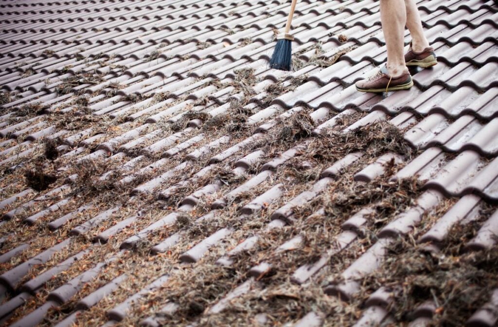 Person standing on a tiled roof, sweeping away piles of dry leaves and debris with a broom. Only their legs and feet are visible.