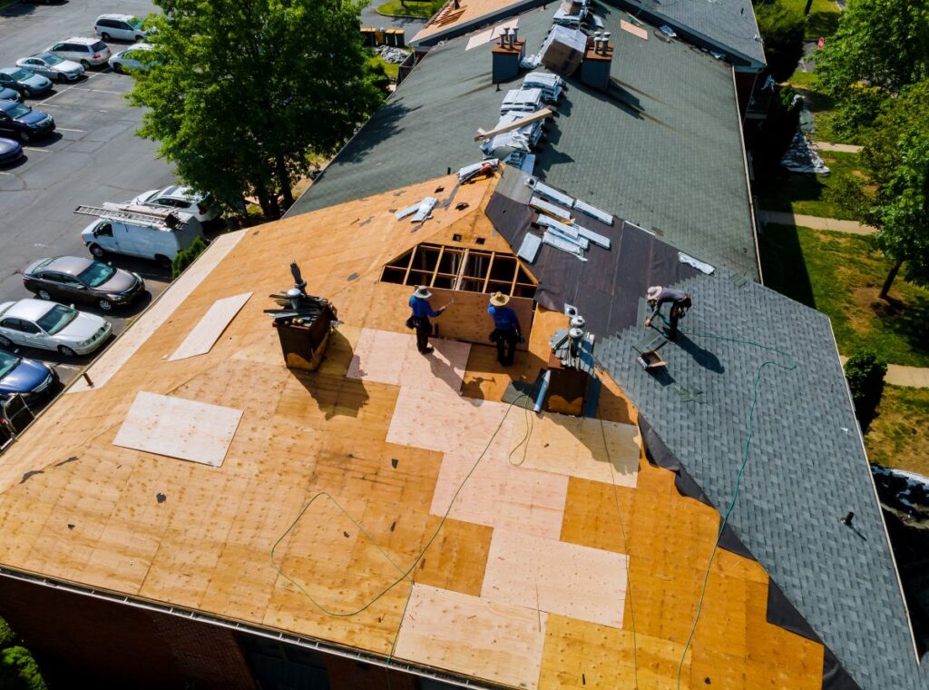 Aerial view of workers repairing or replacing the shingles on a large roof, with part of the roof exposed to wooden panels and construction equipment scattered around. Cars are parked beside the building.