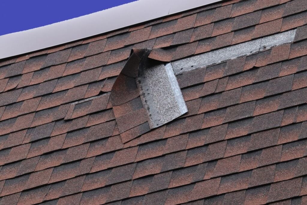 A close-up of a shingle roof with a visible defect where some shingles around a ridge vent are bent and not lying flat, exposing the underlayment beneath. The sky is clear and blue in the background.