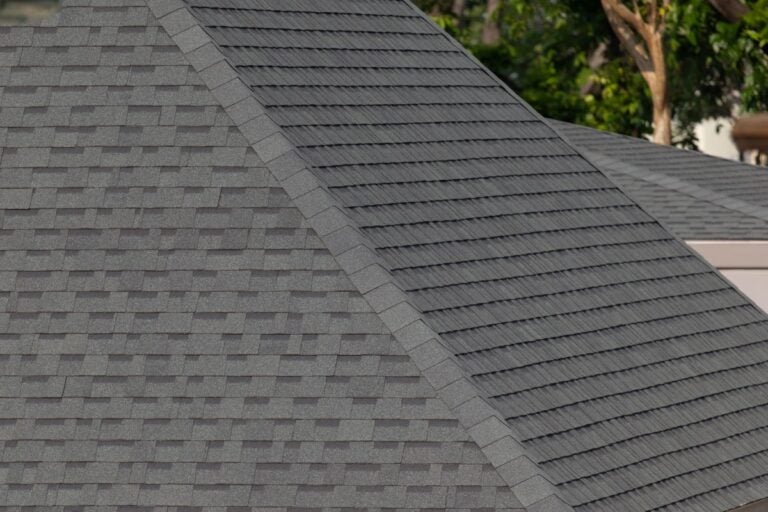 Close-up view of two rooftops with dark gray asphalt shingles, showing the pattern and texture. Trees and part of a building are visible in the blurred background.