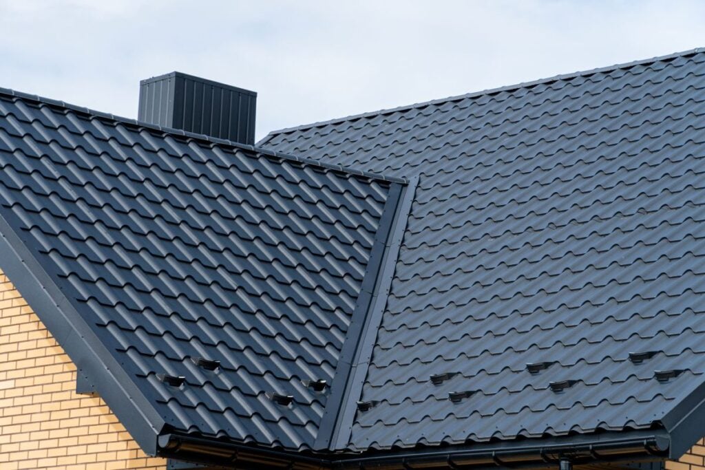 Dark gray metal roof tiles on a modern house with yellow brick walls and a rectangular black chimney, against a cloudy sky.