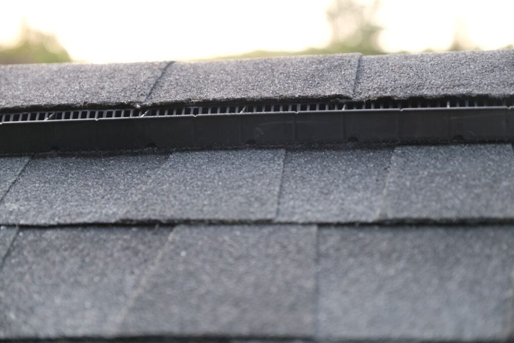 Close-up of a rooftop with dark asphalt shingles and a visible ridge vent, designed to provide attic ventilation. The background is blurred with hints of greenery and sky.