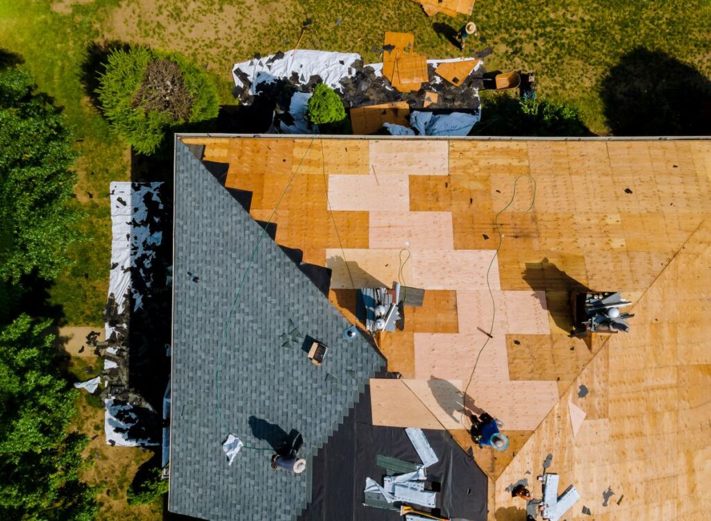 Aerial view of a house roof under repair, with workers installing new shingles and plywood, some areas stripped down and construction materials visible on the ground and roof.