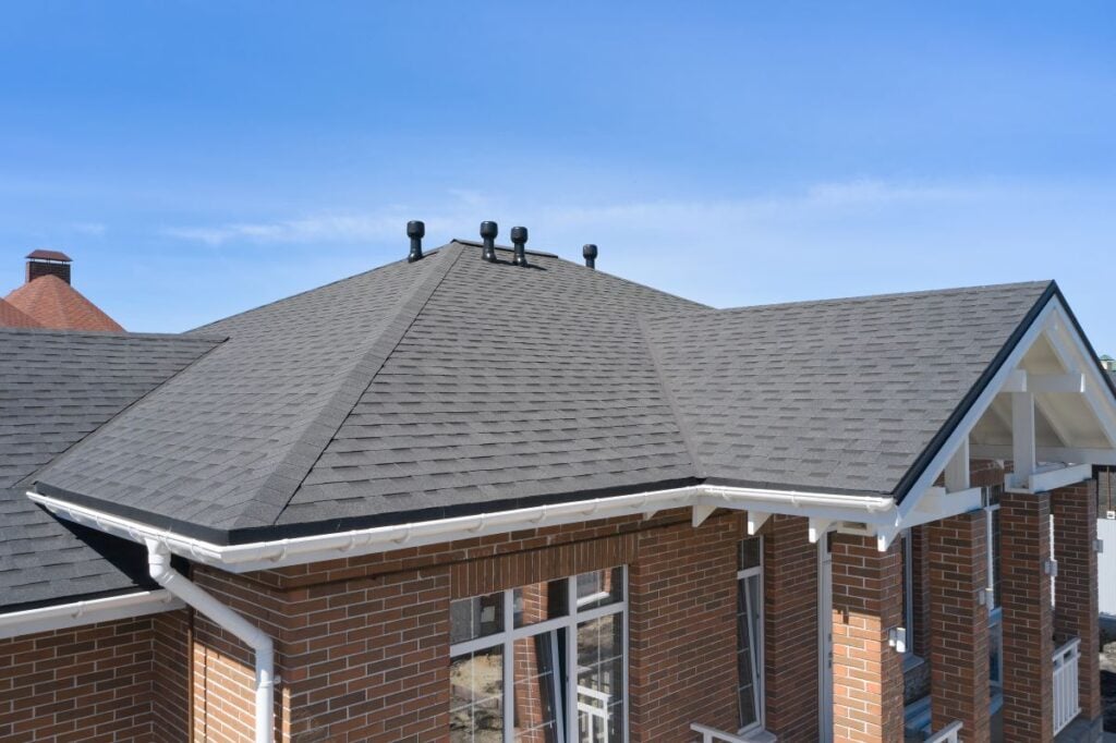 A close-up view of a house roof with dark gray asphalt shingles, several roof vents, white gutters, and a brick exterior under a clear blue sky.