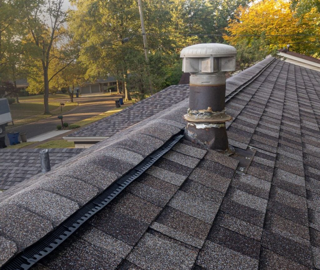 A rooftop with brown shingles, a metal vent pipe, and a black ridge vent. Trees with autumn foliage and a suburban street with houses and trash bins are in the background.