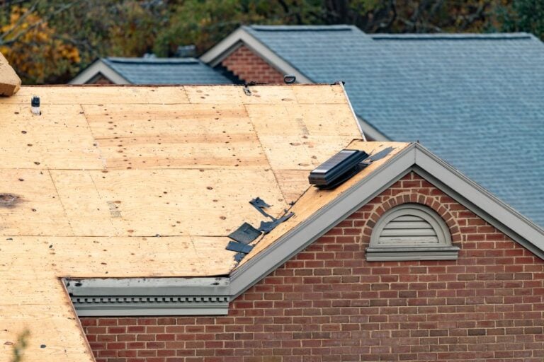 A house with a partially removed roof, showing exposed plywood sheathing, some remaining shingles, and a vent. Neighboring houses with finished gray roofs are visible in the background.