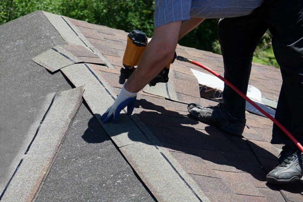 A person wearing a glove uses a nail gun to install asphalt shingles on a house roof, working along the ridge with roofing materials visible nearby.