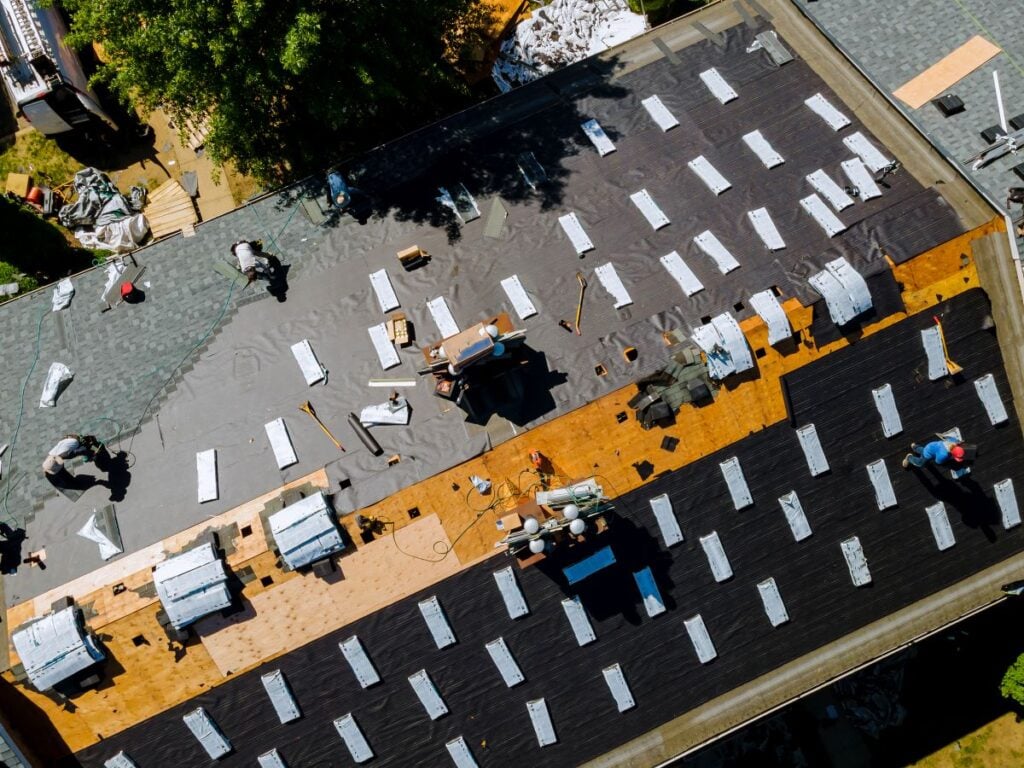 Aerial view of workers installing roofing materials on a large house, with roofing felt, shingles, and tools spread across the roof. Some sections are partially covered, showing progress in the installation process.