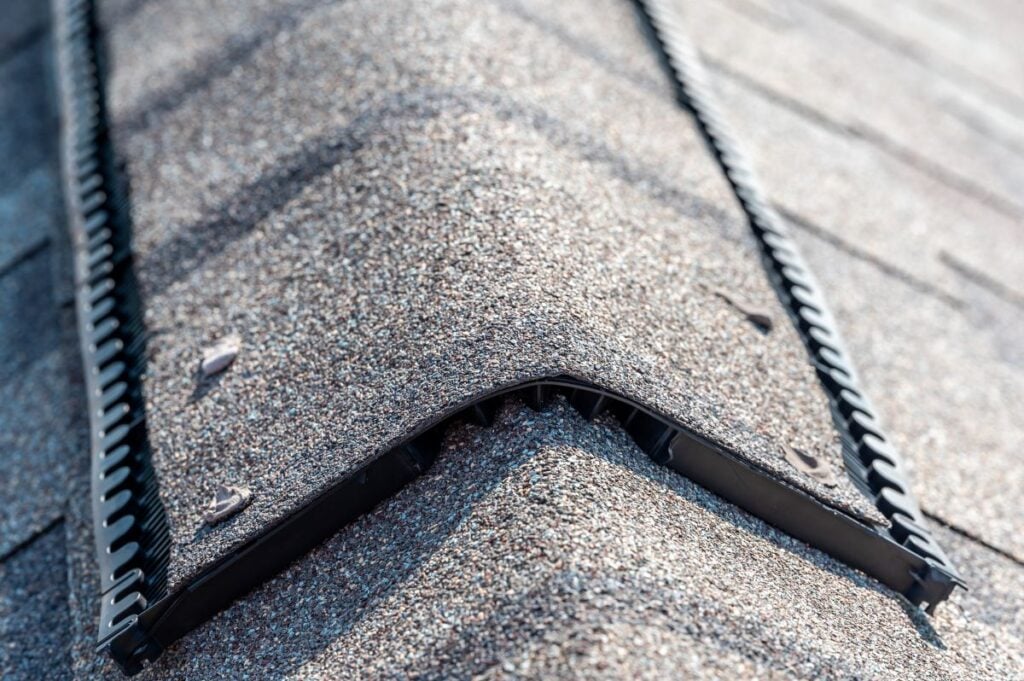 Close-up of a shingled roof ridge with a black plastic ridge vent installed, showing the texture of the shingles and details of the roof ventilation component.